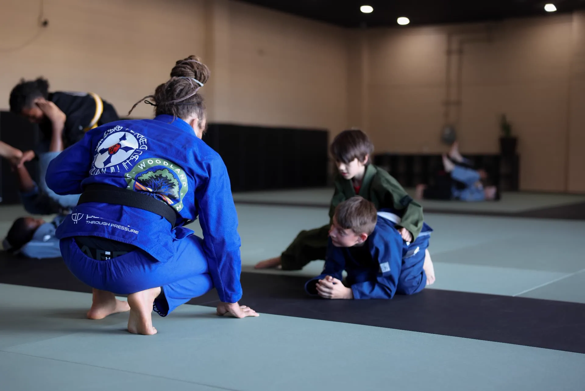 A coach kneeling on the mat guiding two young students through a Jiu-Jitsu drill