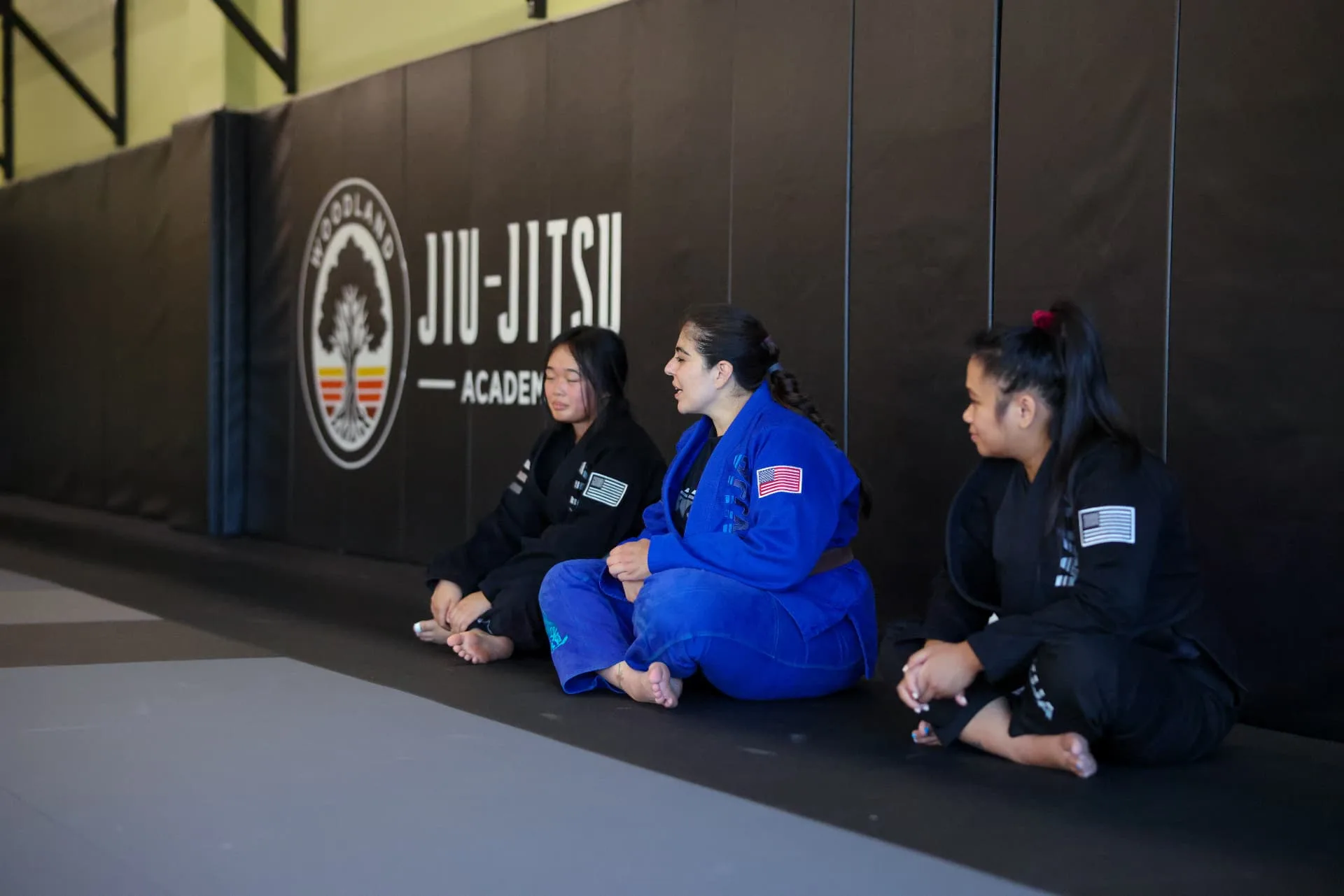 Three women in gis sitting against the wall mats talking through a technique together