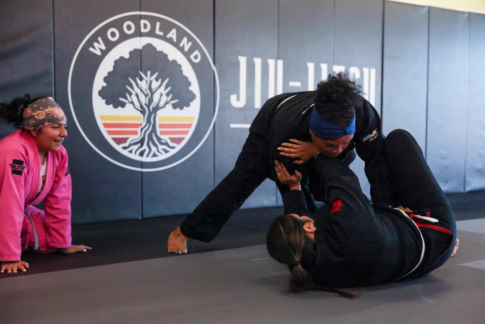 Women training Jiu-Jitsu together on the mat beneath the Woodland Jiu-Jitsu wall logo