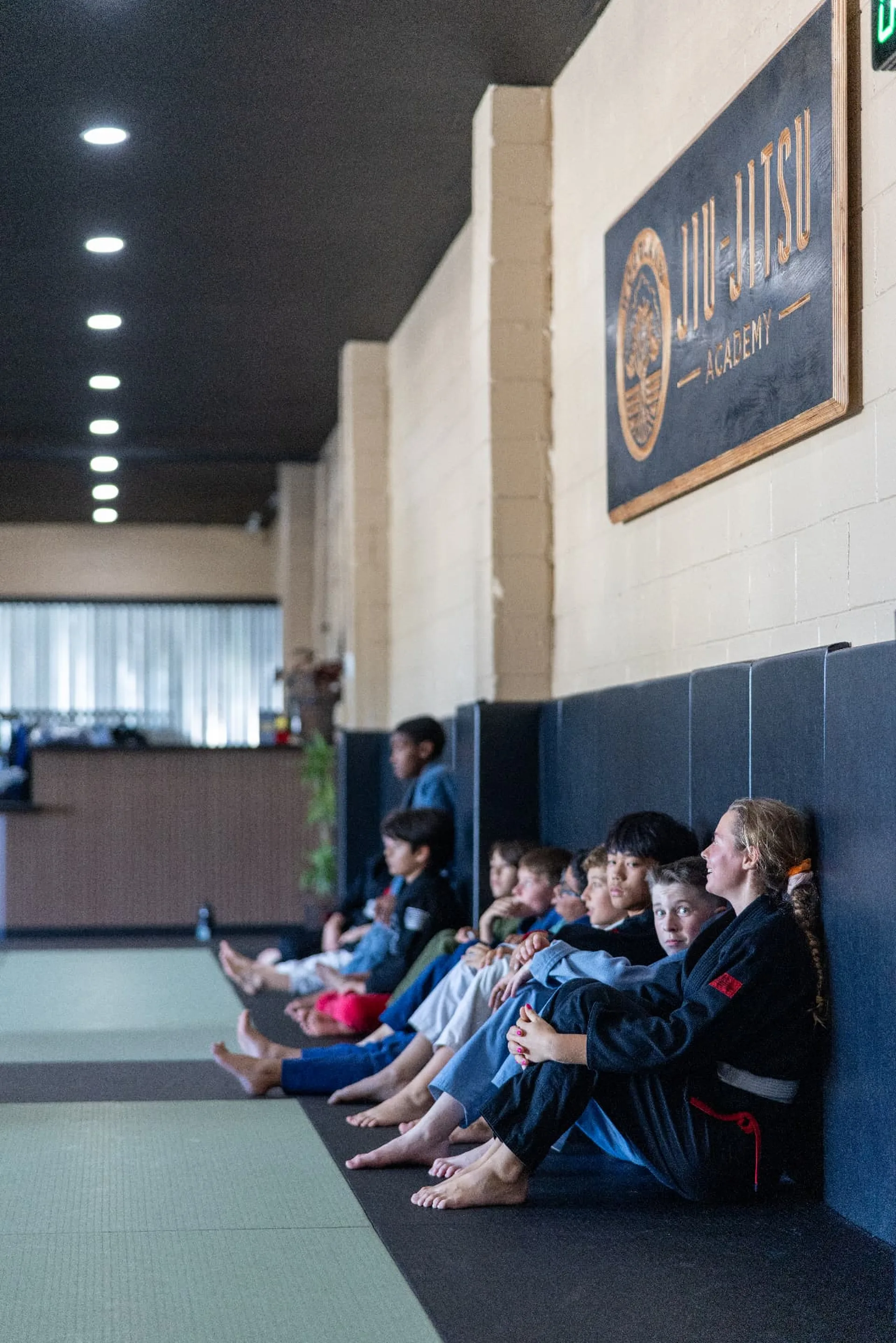 A row of kids in gis sitting along the wall watching a technique demonstration