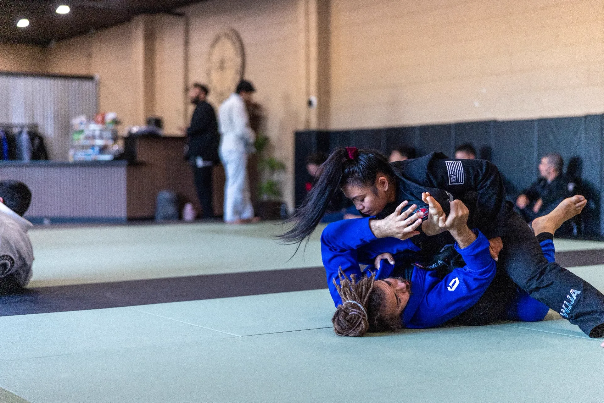 A coach in a blue gi working from guard against a student in a black gi during adult sparring