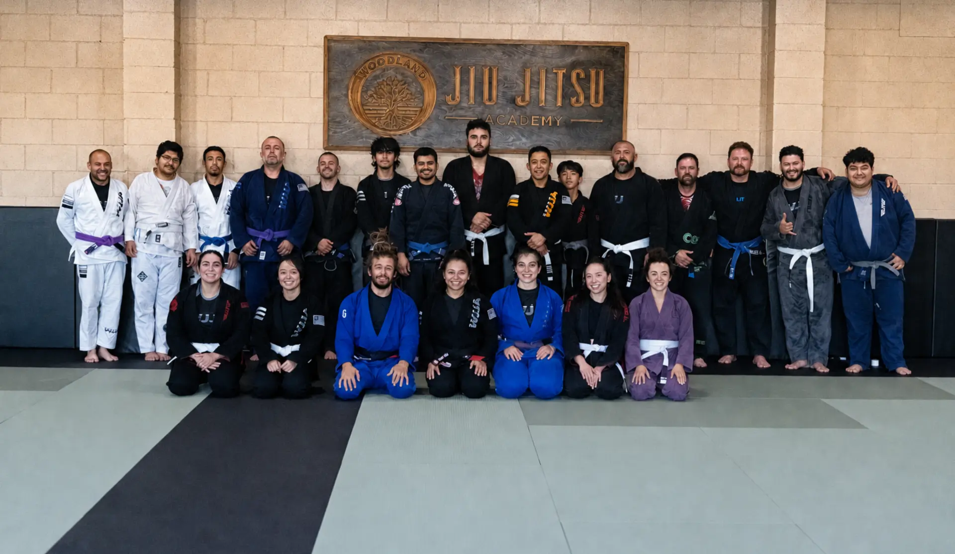 The full adult class at Woodland Jiu-Jitsu Academy gathered on the mat in their gis beneath the carved academy sign after training