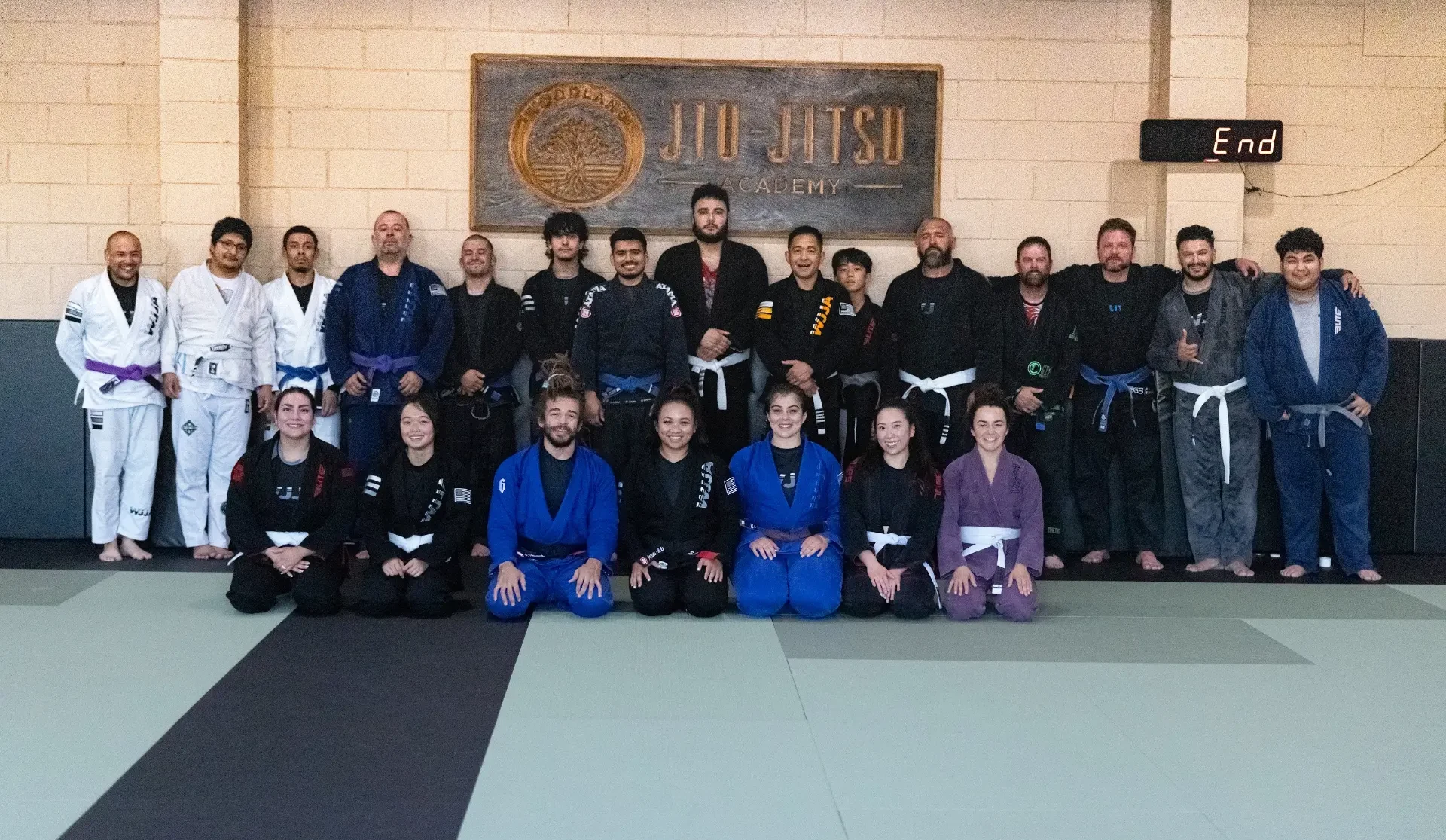 The full adult class at Woodland Jiu-Jitsu Academy gathered on the mat in their gis beneath the carved academy sign after training